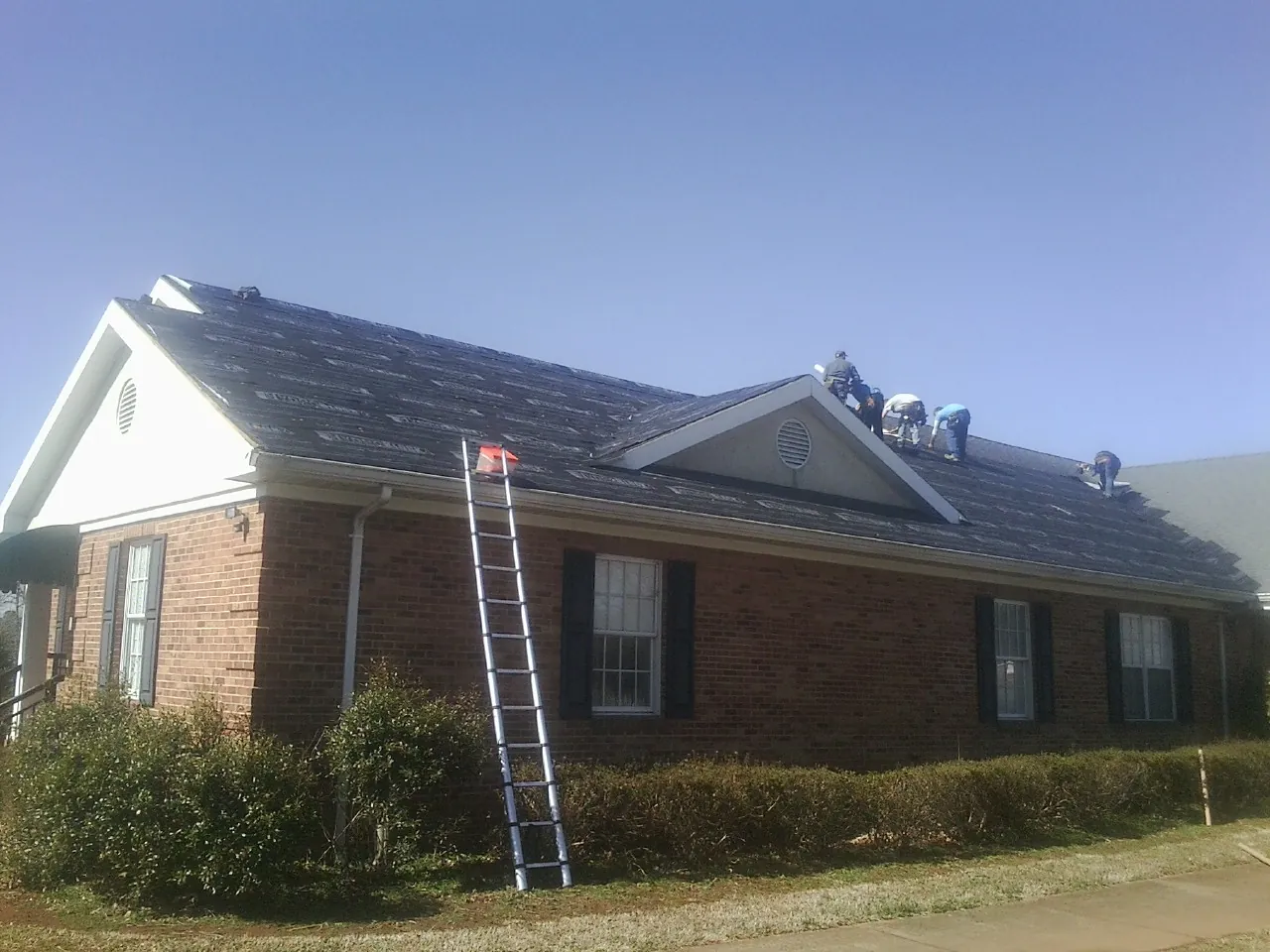 Crew tearing shingles off a brick ranch roof with a ladder set — Metal Roof Repair work by our crew in Connelly