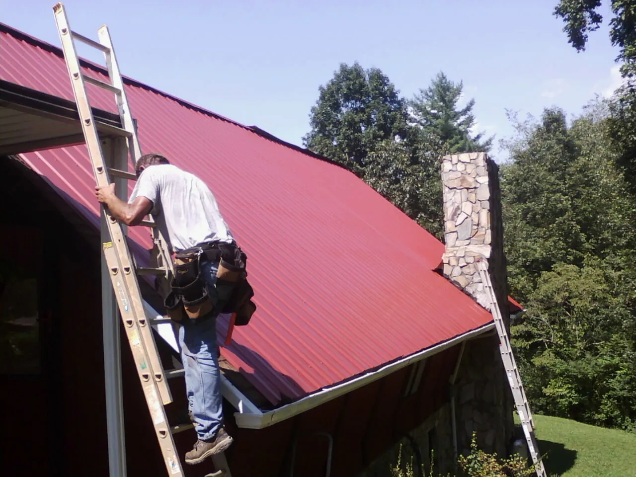 Roofer on a ladder beside a red metal roof and stone chimney — Soffit & Fascia Repair by Roofing Brothers in Connelly